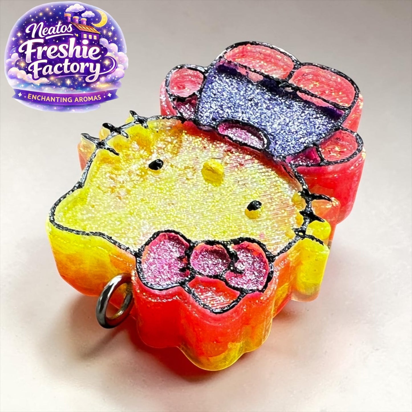 Neatos Elements Island Sunset Hello Kitty car freshie, yellow face, pink bow, red and blue accents, glitter details.