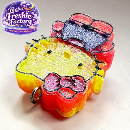 Neatos Elements Island Sunset Hello Kitty car freshie, yellow face, pink bow, red and blue accents, glitter details.